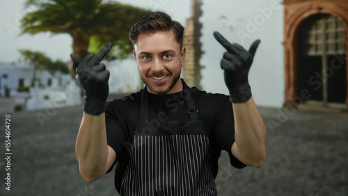 Young man with beard gestures humorously with middle fingers on a city street while wearing black gloves and a striped apron, appearing amused in an outdoor urban setting.