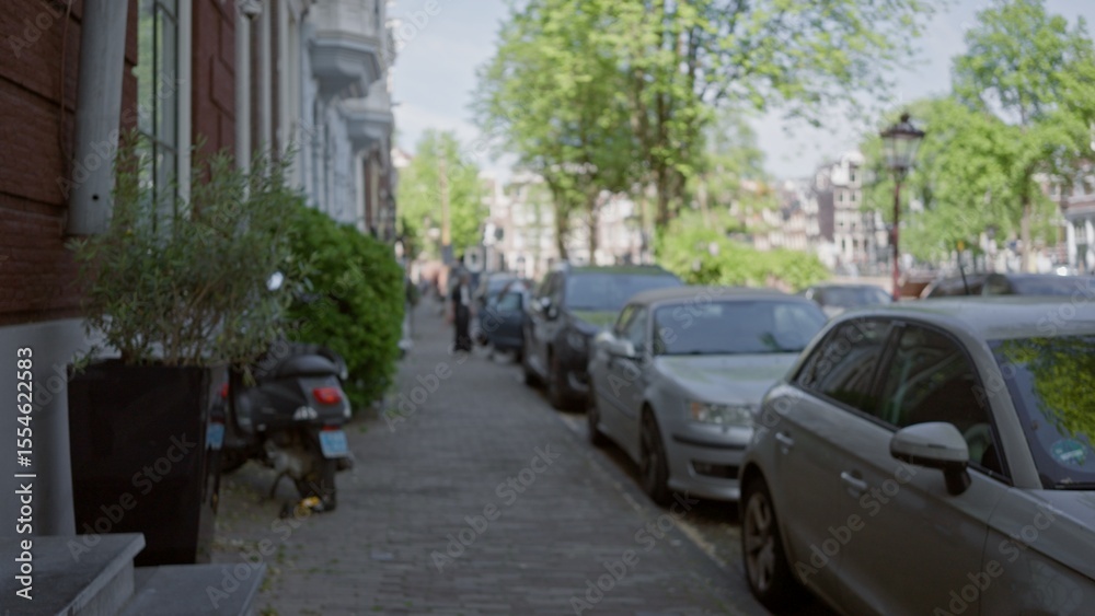 Obraz premium Blurred city street scene in a european town with parked cars, leafy trees, and people walking on a sunny day.