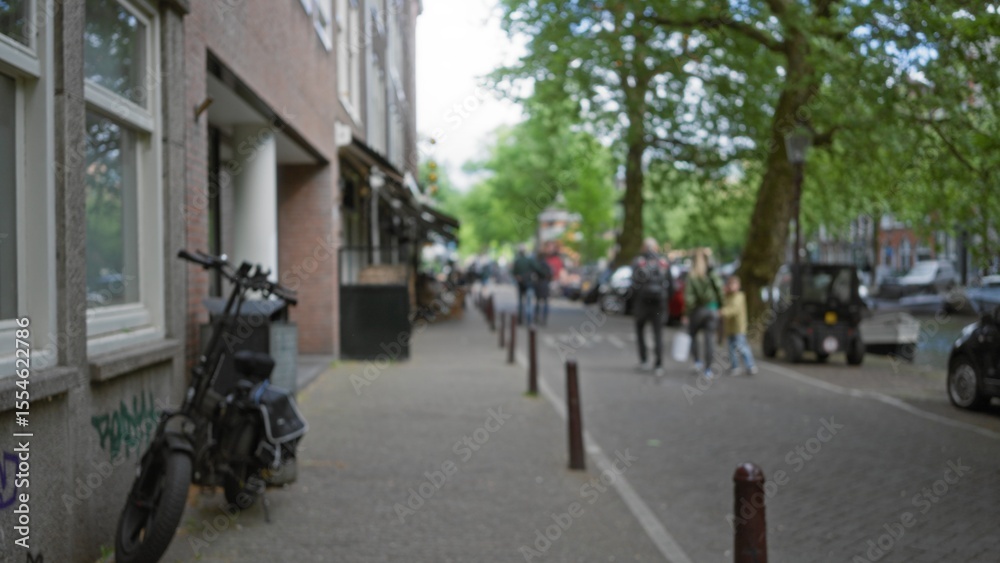 Obraz premium Blurred street scene with people walking on a european city sidewalk, showcasing urban life, bokeh effect, and defocused background, trees and bicycles visible.