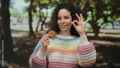 Young hispanic woman wearing colorful sweater in park makes ok gesture while enjoying delicious cookie in an outdoor setting filled with trees and sunlight.