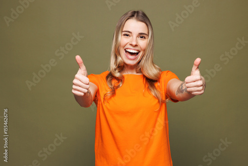 Young smiling happy Caucasian woman she wears orange t-shirt casual clothes looking camera showing thumb up like gesture isolated on plain pastel green background studio portrait. Lifestyle concept.