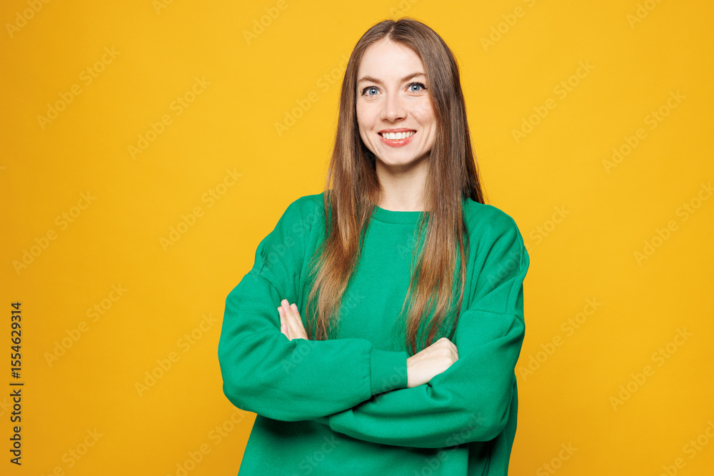 Fototapeta premium Young smiling happy fun Caucasian woman she wears green sweatshirt casual clothes hold hands crossed folded look camera isolated on plain yellow orange background studio portrait. Lifestyle concept.