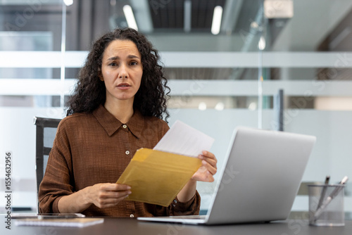 Fényképezés A concerned businesswoman in her office is holding documents, sitting in front of a laptop, looking directly at the camera