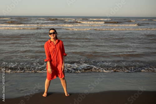 Happy woman in a red dress enjoys a peaceful beach walk at sunset, radiating joy and freedom during a relaxing seaside vacation. Travel moments full of emotion