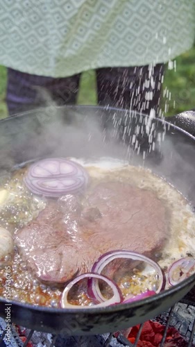 Vertical video. Food preparation. Chef adding salt to steak. Juicy Steak cooking in a cast iron pan on a grill outdoors. Slow motion footage 100fps.