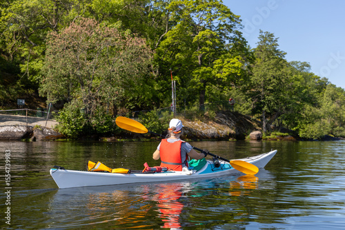Konstfotografi Woman paddling a white kayak along a calm, green shoreline