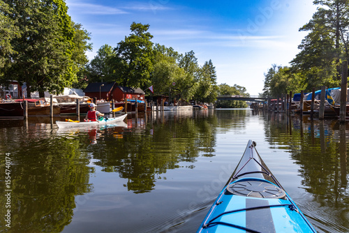 Quadro em tela Two kayaks moving through a quiet canal in central Stockholm, surrounded by moored boats, red boathouses and leafy trees