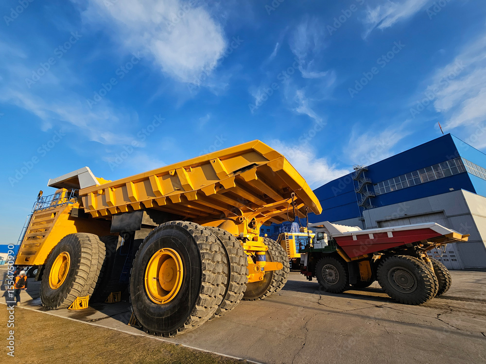 Fototapeta premium A large yellow dump truck is parked next to a building. The sky is blue and there are clouds in the background
