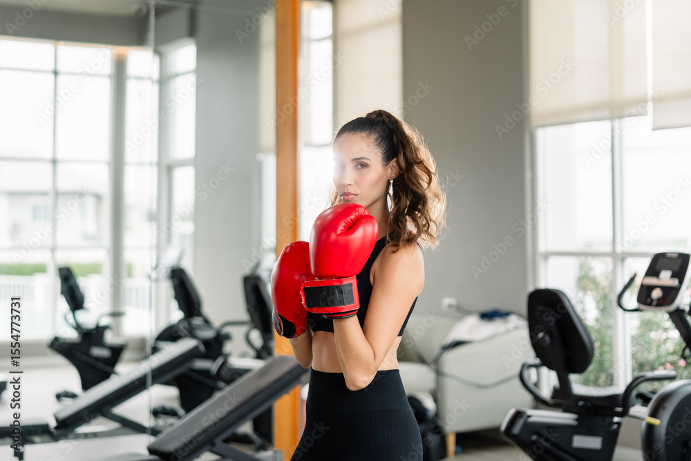 Fototapeta premium A woman wearing a red boxing glove stands in a gym. She is wearing a black tank top and black pants