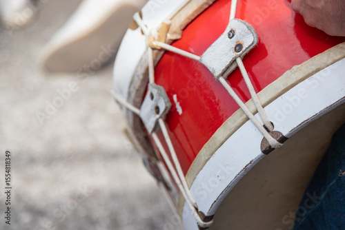 Close-up of a person playing a red and white rope-tension bass drum during an outdoor cultural event, capturing traditional percussion details and rhythmic performance energy.