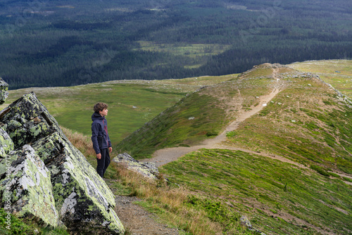 A young boy stands on a rocky ledge of Städjan mountain, gazing out over the lush green landscape of Idre, Sweden, while enjoying nature on his vacation