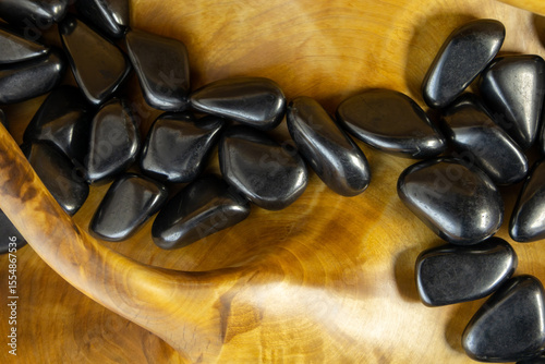 Wooden bowl filled with shiny black stones sits on a wooden table. The bowl is curved and has a unique shape, making it an interesting and eye-catching display. The stones are of various sizes