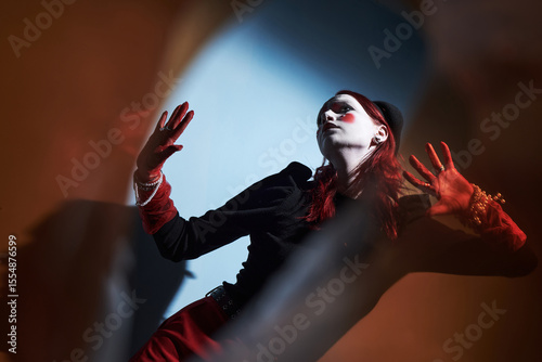 Caucasian teenager girl with creative makeup and red hair performing expressive dance gesture at home, hands raised, wearing jewelry, embodying artistic Gen Z style