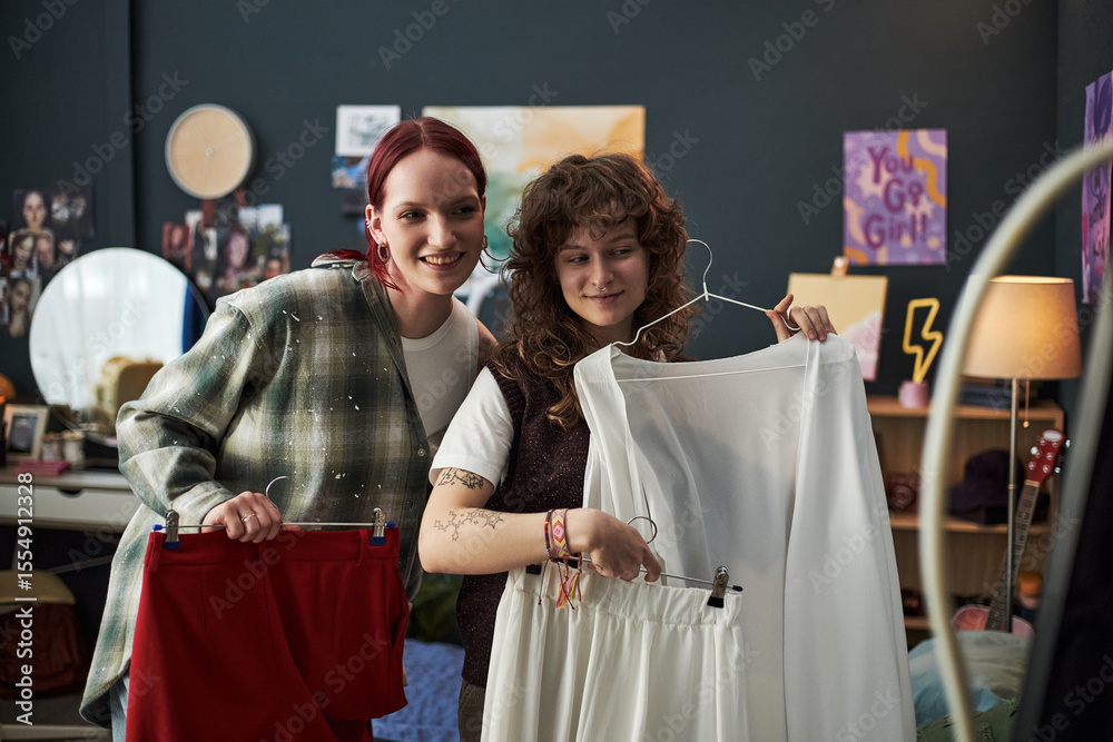 Obraz premium Two Caucasian teenage girls smiling while holding up clothing in bedroom, one with red hair and one with curly brown hair, engaging in creative fashion activity at home together