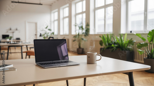 Laptop and coffee mug on a desk in a bright office with plants and large windows in the background