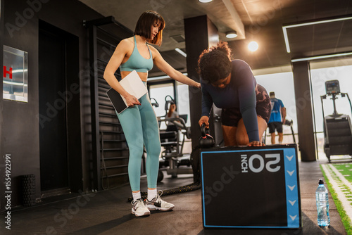 Overweight woman is doing exercises with a kettlebell in a gym, guided by her personal trainer, who is holding a tablet and providing instructions and support