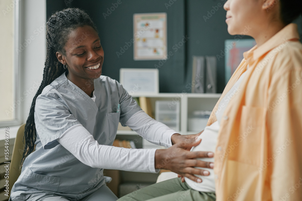 Obraz premium Black woman healthcare professional examining pregnant Asian woman during prenatal care checkup in medical office, focusing on abdominal assessment and patient wellbeing