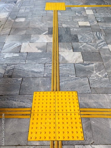 textured yellow markings guide visually impaired individuals across tiled walkway. sunny location features contrasting surfaces for better accessibility. close up.