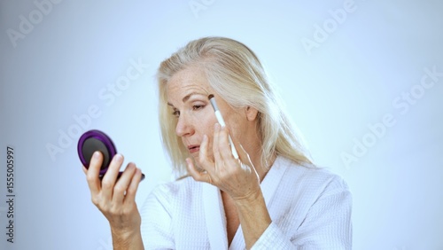 A mature woman in a white robe applies makeup with a brush, enjoying the relaxing atmosphere of the spa. Her joyful expression reflects her self-care moment.