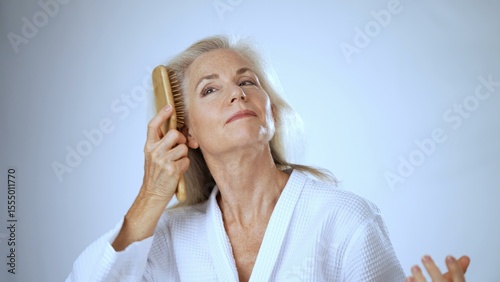 A happy mature woman in a white robe gently runs her fingers through her long hair, enjoying a moment of relaxation at a peaceful spa environment.