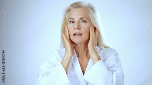 A mature woman in a white robe expresses happiness while at a spa. Her hands gently touch her neck as she basks in the soothing atmosphere, radiating positivity.