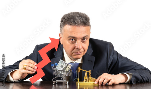 A middle-aged man in a business suit holds a red arrow of a financial growth chart in his hand, a gold oil pump and a $100 bill in a miniature shopping cart are on the table in front of him