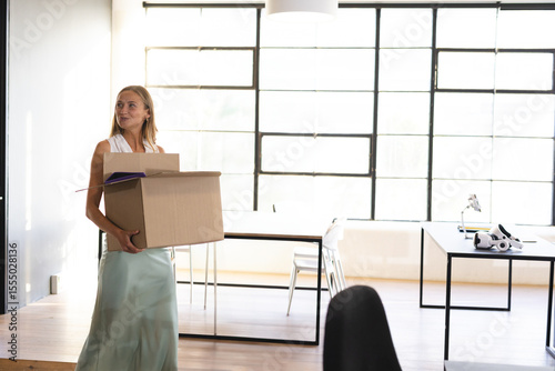 Woman carrying cardboard box with folders in open-plan office with VR headset on desk, copy space