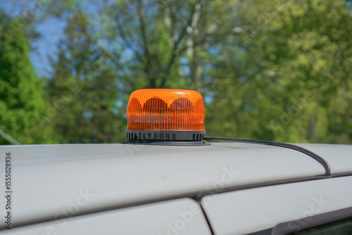An orange warning beacon light with a black base and wire mounted on the white roof of a vehicle, with blurred green trees in the background, concept of emergency services, construction, road safety