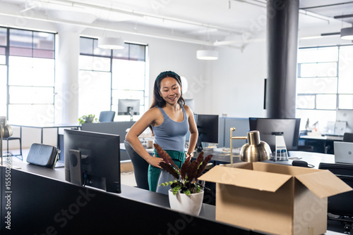 Tableau sur toile Asian Chinese woman standing unpacking items at open-plan office desk with monit