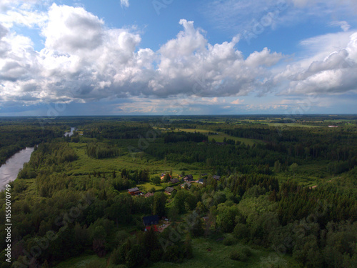 Aerial view of a peaceful rural landscape in Tartu County, Estonia. A small village, green forests, fields, and a river under a partly cloudy sky. Scenic summer countryside.

