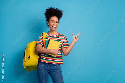 Cheerful young woman in striped shirt with backpack holds book and points excitedly on a light teal background
