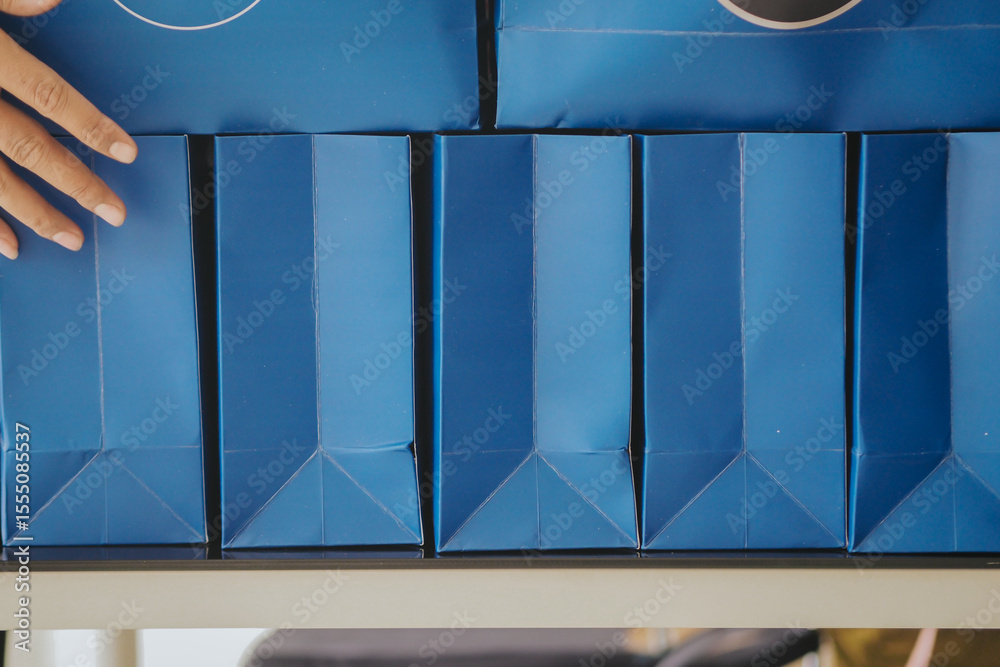 Ταπετσαρία hand reaching over several identically sized and shaped dark blue gift bags or boxes, neatly lined up in a row on a surface
