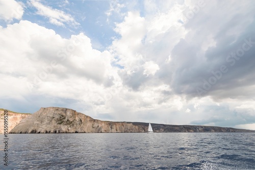 A sailboat navigates smoothly on tranquil waters, surrounded by towering cliffs beneath an expansive sky filled with fluffy clouds. This coastal landscape captures nature’s beauty at midday