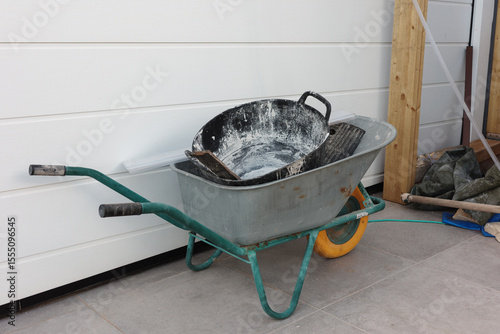 A used construction wheelbarrow is filled with a plastering tray and hand tools, standing on a tiled surface near a white wall. Concept of renovation, manual labor and outdoor construction process.