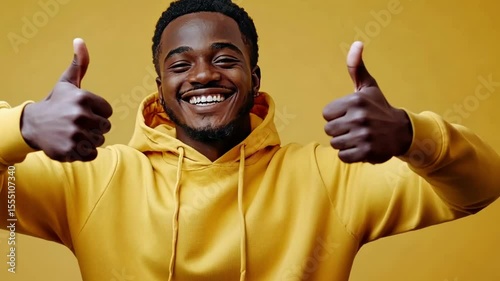 Cheerful young man gives thumbs up while wearing a yellow hoodie against a bright background