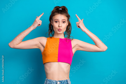 Young woman with a colorful top pointing upwards humorously against a blue backdrop, showcasing playful style
