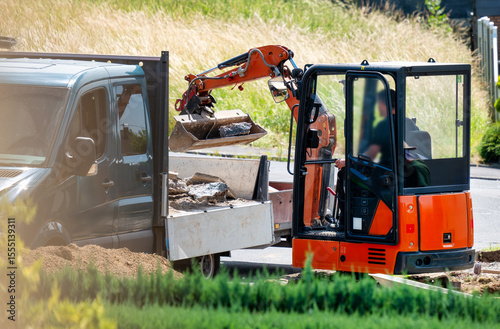 A mini excavator loads rubble into a flatbed truck during landscaping or construction work