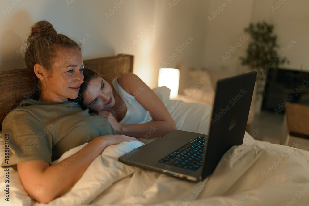 Fototapeta premium Middle age woman and daughter relaxing together in bed with laptop, enjoying wellbeing, healthy lifestyle, and warm family moments at home in cozy lighting.