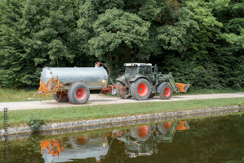 Farming activity near a reflective pond with a tractor and tank in a lush green countryside setting