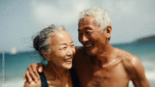 Happy elderly Asian couple embraces while laughing on a sunny beach
