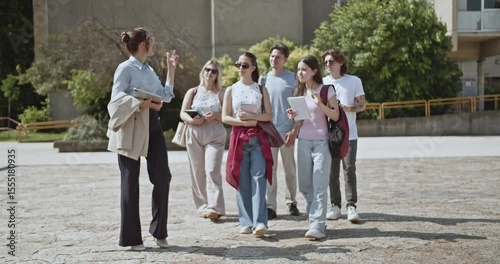 Female teacher leads a diverse group of young university students on a guided tour across the campus grounds, explaining and sharing educational insights. They are learning and engaging in discussion.