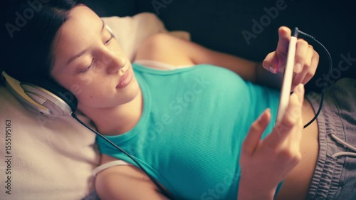 A serene and intimate close-up of a young woman relaxing, lying down with headphones on, engrossed in content on her smartphone. Ideal for themes of relaxation, digital leisure, music, communication.