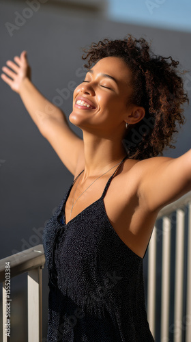 Young African American woman stretching out her arms joyfully in the morning sunlight on a balcony. 