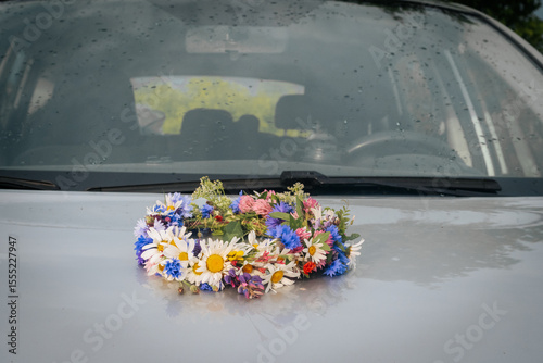 A colorful wreath of wildflowers rests on the wet hood of a car, reflecting the light of a recent rain. The windshield and wipers are visible in the background.