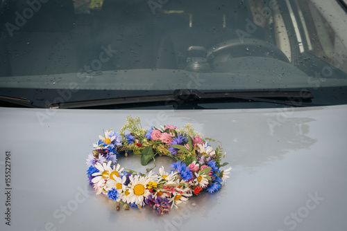 A colorful wreath of wildflowers rests on the wet hood of a car, reflecting the light of a recent rain. The windshield and wipers are visible in the background.