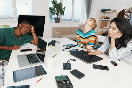 Tired woman and her colleagues socially disengaged at workplace, young adult coworkers from Gen Z in casual work environment socializing and working together.