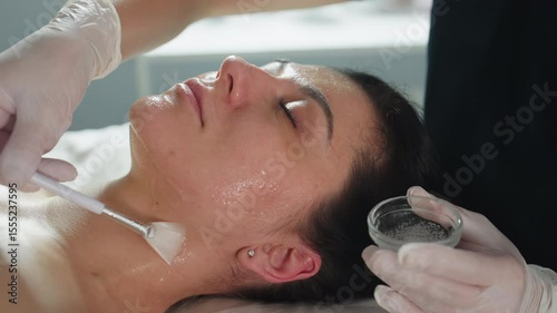 Female groomer in gloves applies clear facial massage gel with brush from chin up to neck on relaxed client lying on spa bed under soft clinical light for nourishing skincare treatment session