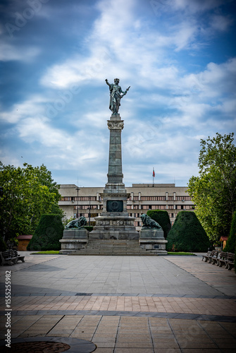 Monument of Liberty in Rousse, Bulgaria Under a Cloudy Sky. Prominent sculpture features a female figure atop a tall stone column, flanked by bronze lions, representing national freedom and resilience