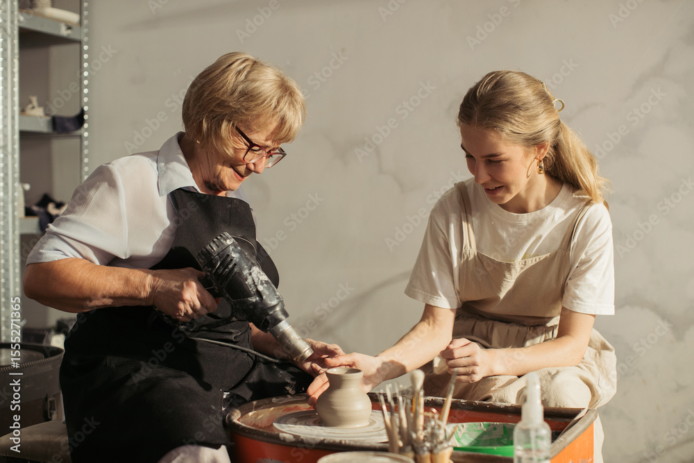 Fototapeta premium Senior potter drying clay pot with heat gun while teaching apprentice