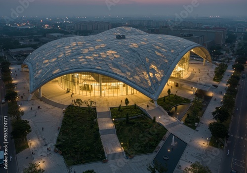 Aerial night view of an illuminated modern architectural building with a unique geometric roof and surrounding landscape design, showcasing urban elegance.